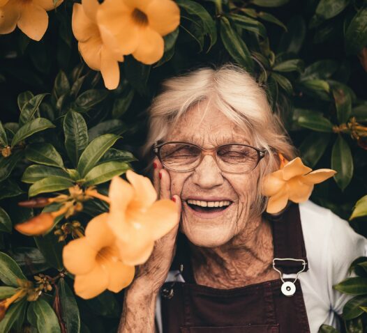 En äldre kvinna ler omgiven av gröna blad och stora orange blommor.